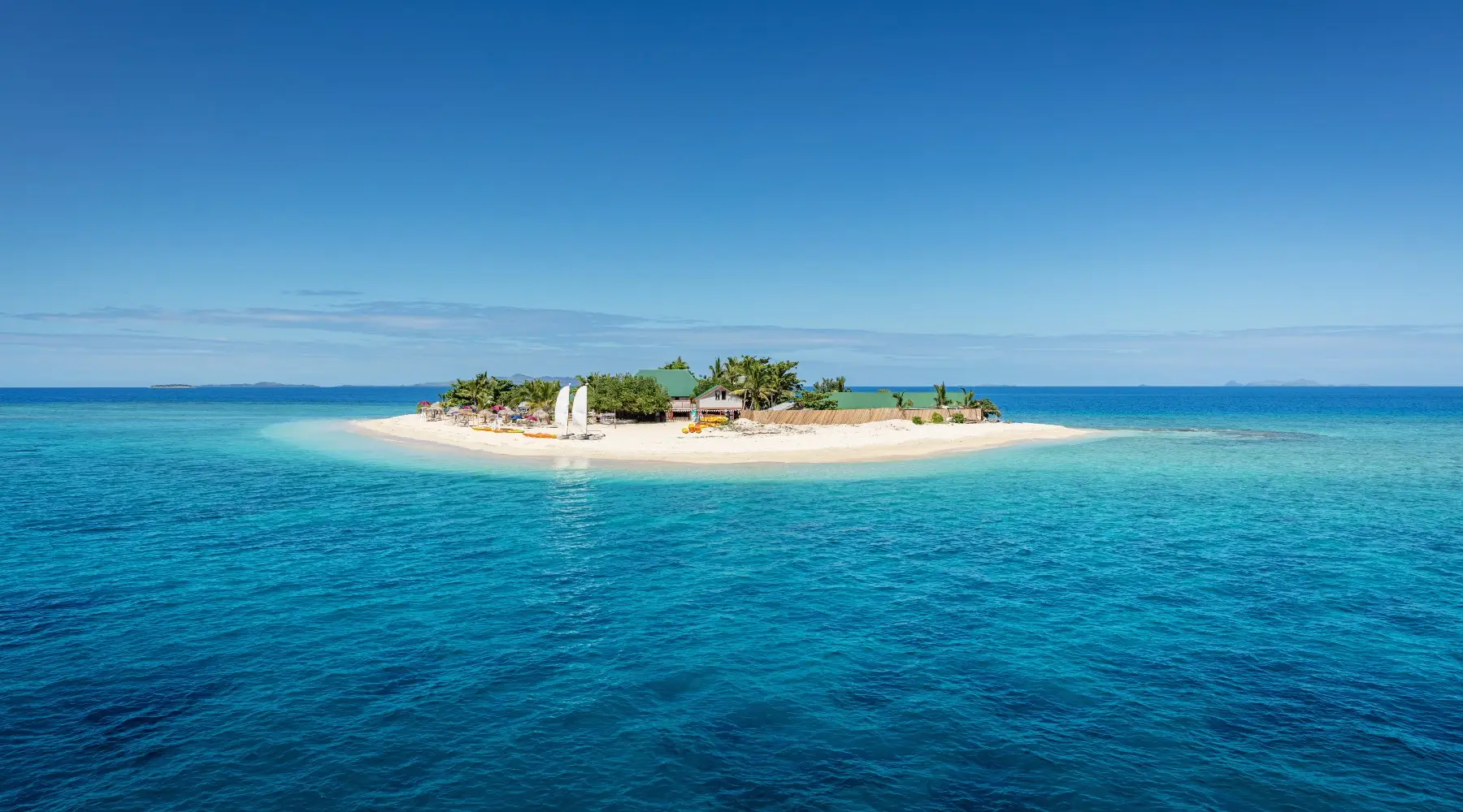 Petite île au milieu de l'océan pacifique sud avec cabines de plage, chaises longues, palmiers, entourée d'une eau turquoise claire.