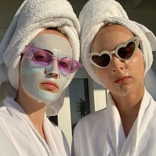 Selfie of two girls with their hair wrapped up in white towels, wearing sheet masks on their face and coloured sunglasses on top
