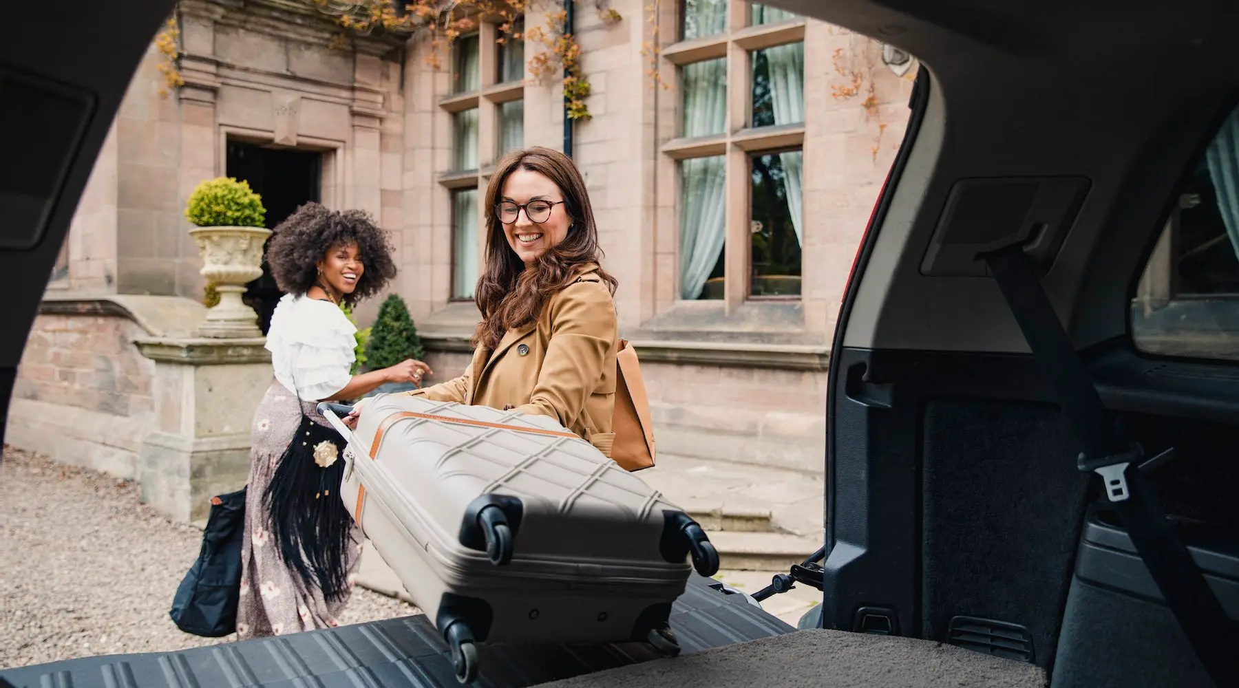 Duas mulheres puxando malas de um carro, com um belo prédio histórico atrás delas.