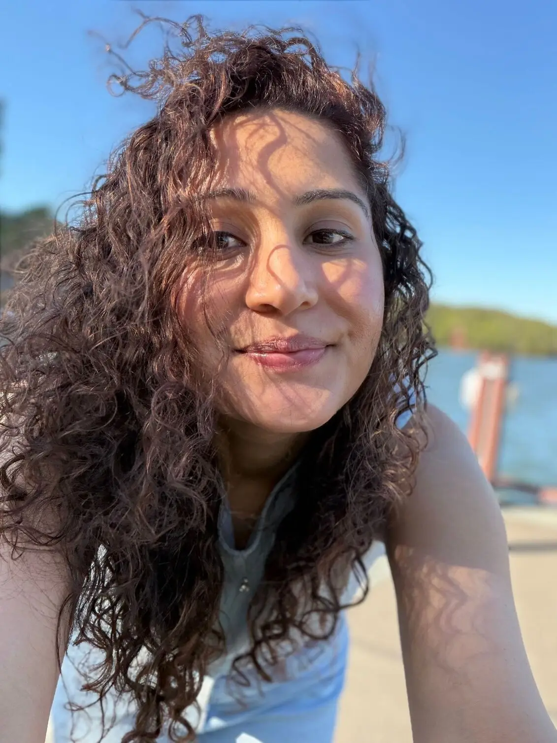 Photo using iPhone 17 close-up selfie of a woman smiling brightly in direct sunlight outdoors.