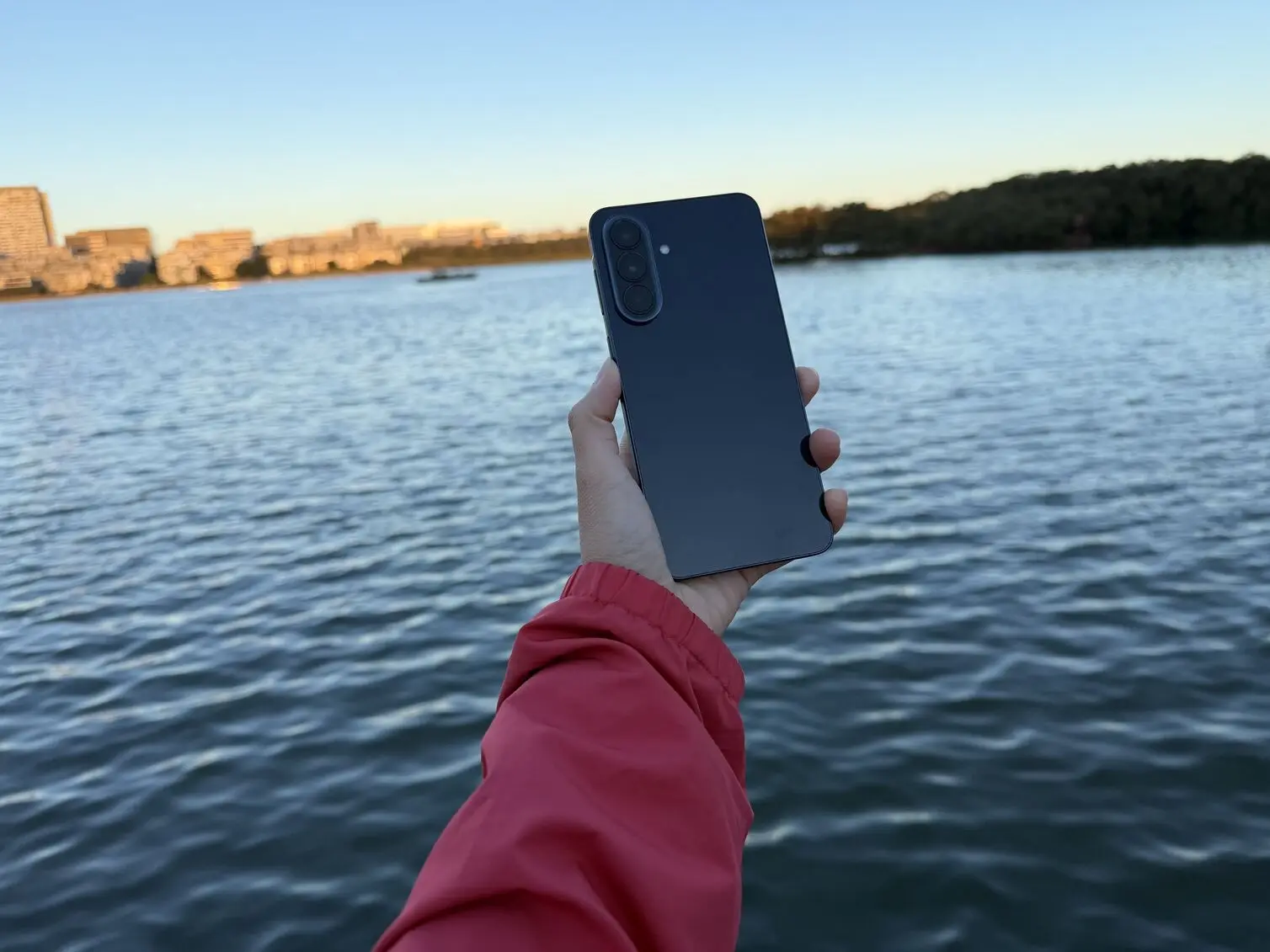 A person holding Samsung Galaxy A57 that displays the back features of the device against the backdrop of a calm river.