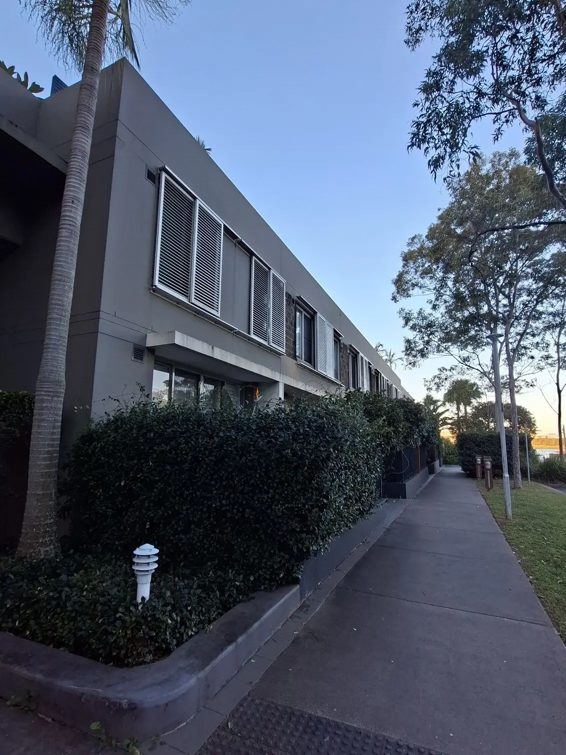 A concrete footpath runs alongside a modern grey apartment building lined with green hedges and tall trees image taken using the Samsung Galaxy A57.
