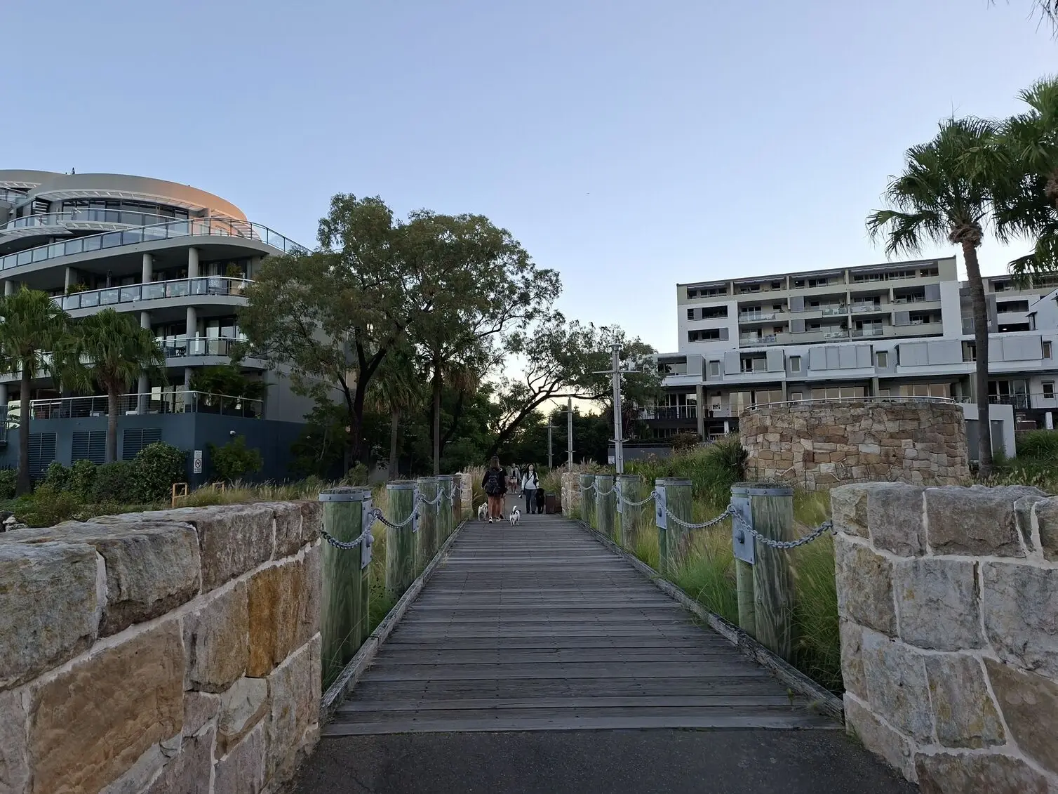 A wooden boardwalk flanked by stone walls leads towards people walking dogs between modern apartment buildings image taken using the Samsung Galaxy A57.