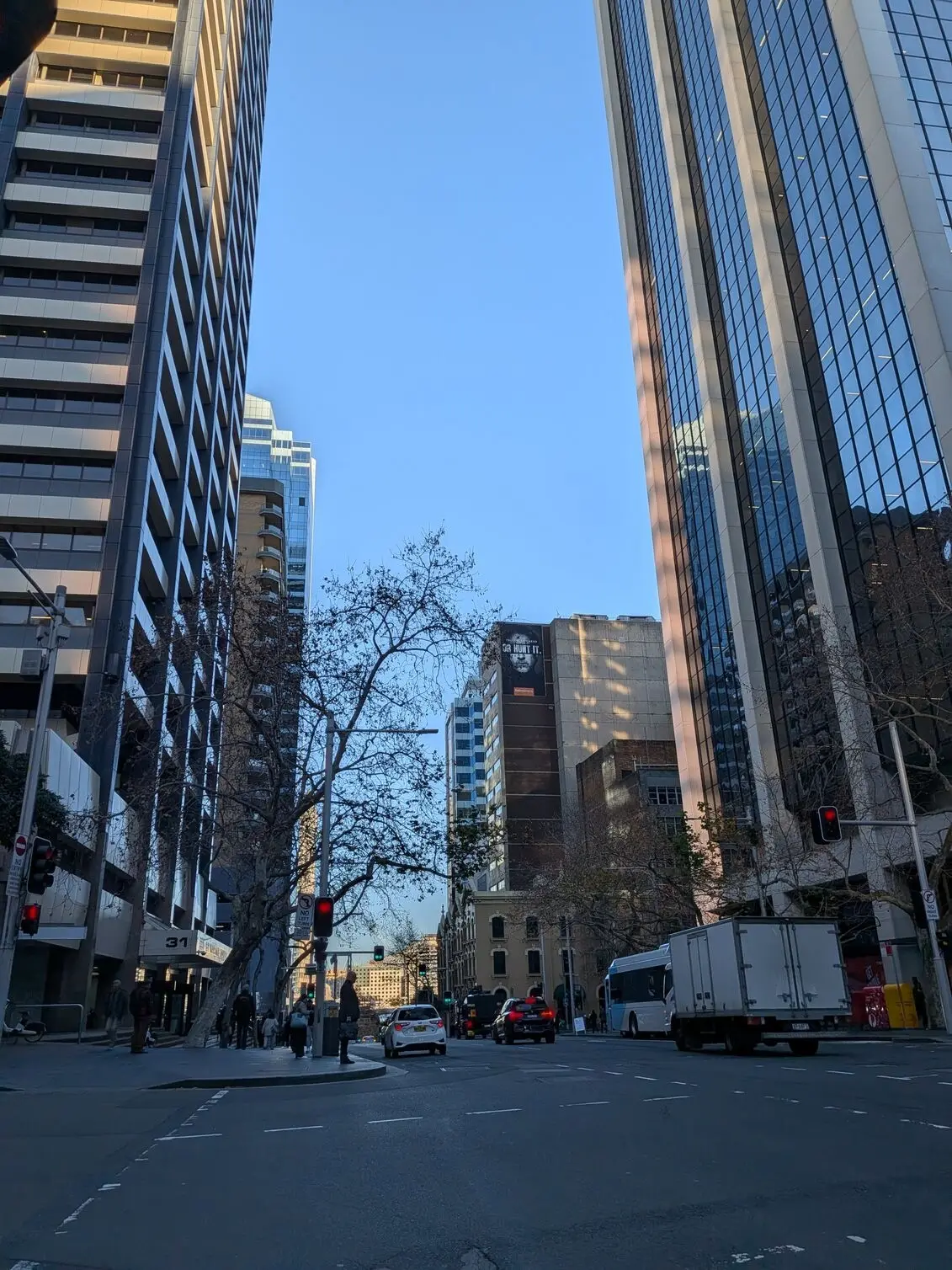 A dramatic upward-angled cityscape shot on the Google Pixel 10a, framed by towering skyscrapers along the street.