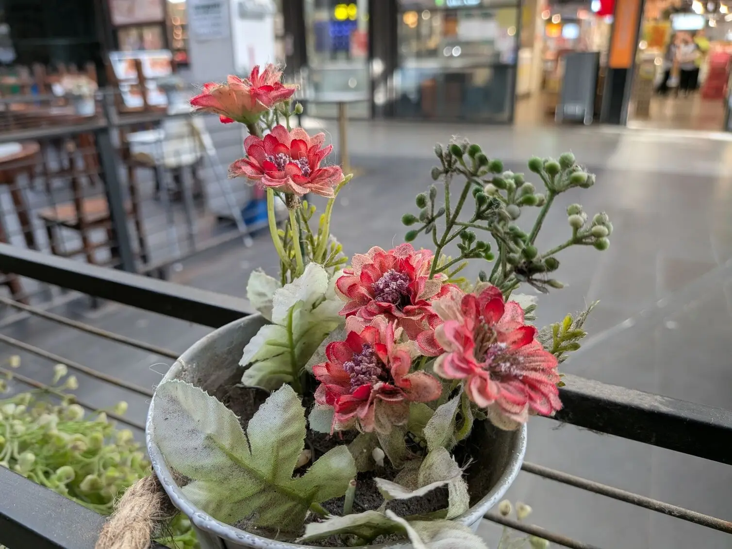 A close-up shot using Google Pixel 10a of an artificial floral arrangement in a rustic pot, with a blurred shopping mall background showcasing the camera's depth-of-field capabilities.