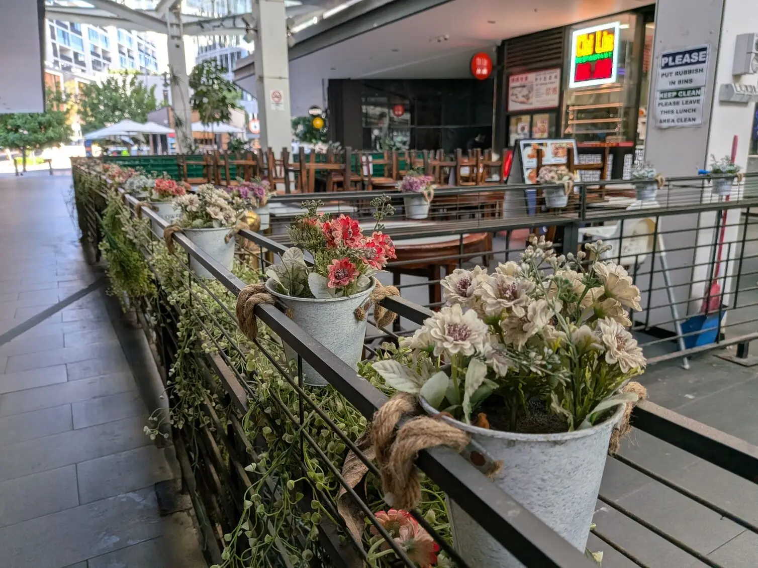 Google Pixel 10a photographs a row of potted flowers hanging along a railing in an outdoor area.