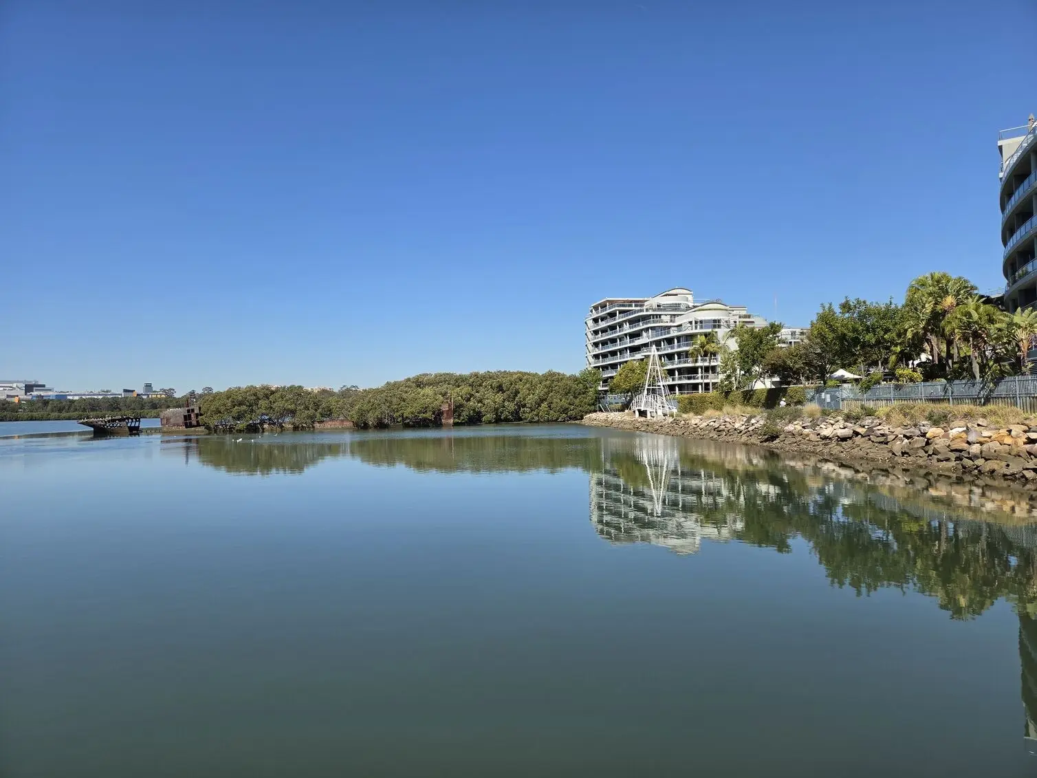 A still river shot on the Samsung Galaxy Z Fold 8, capturing mirror-like reflections of nearby apartment buildings and a distant shipwreck. 