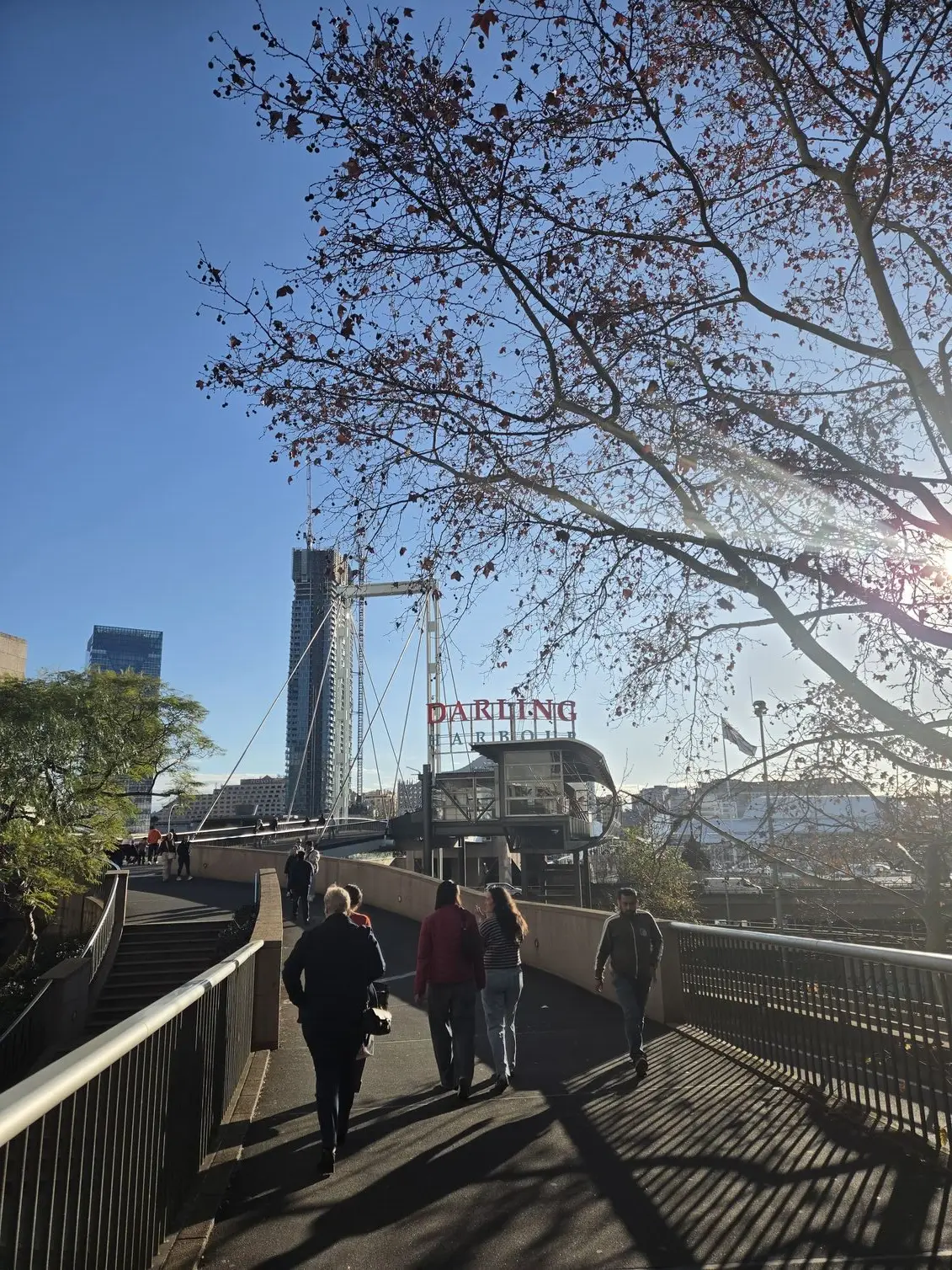 A wide street scene near the footbridge, taken on the Samsung Galaxy Z Fold 8 to show its handling of overhead sun glare and layered depth. 